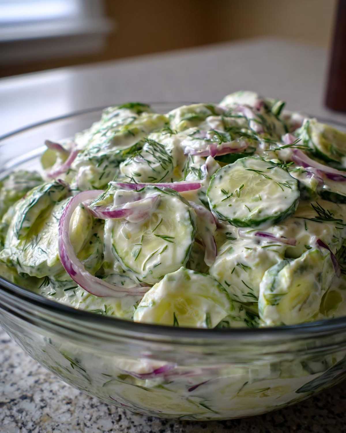 Close-up of a large glass bowl filled with creamy cucumber salad, featuring sliced cucumbers, red onions, and fresh dill.