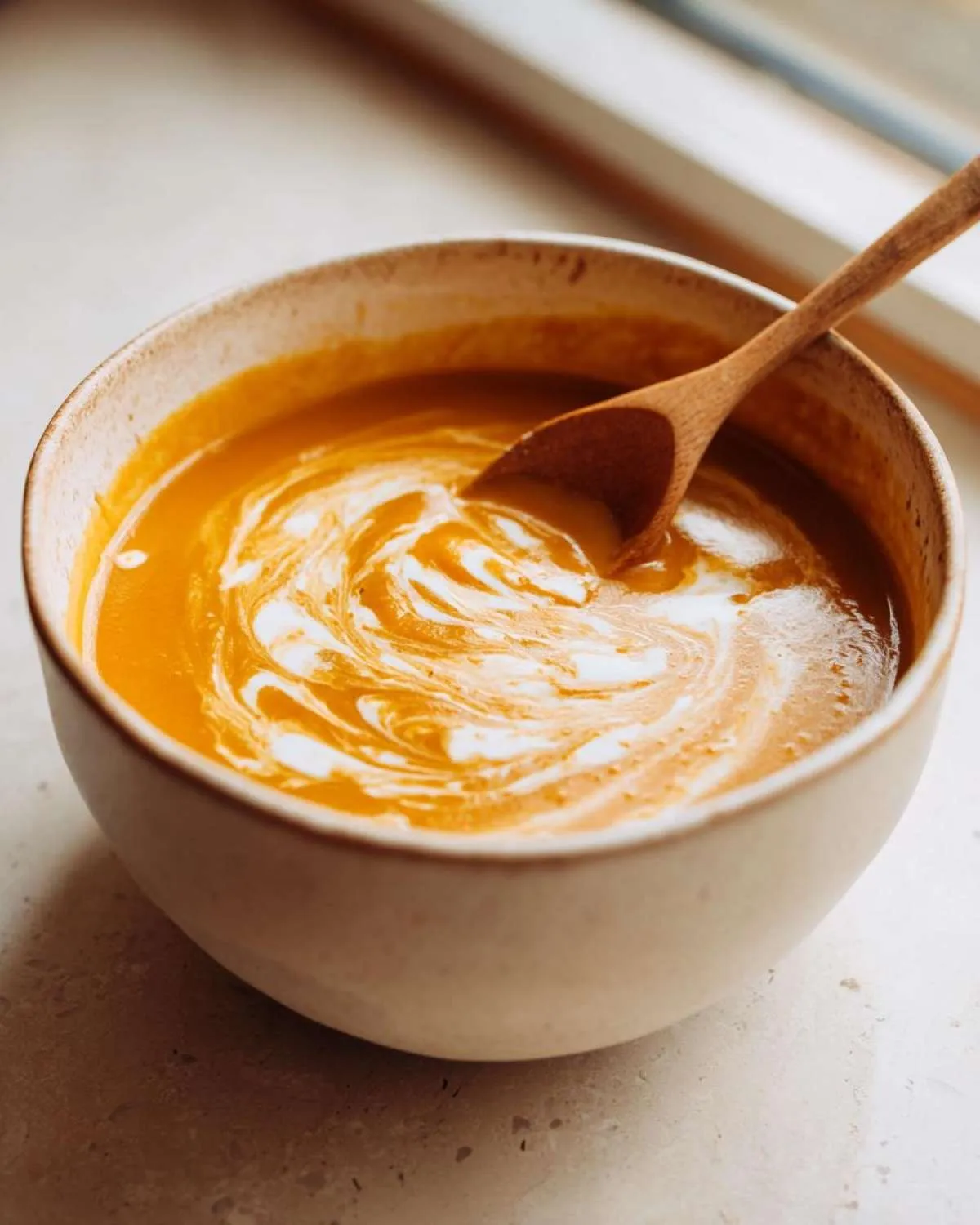 A bowl of creamy fall soup with a swirl of cream and a wooden spoon resting inside.