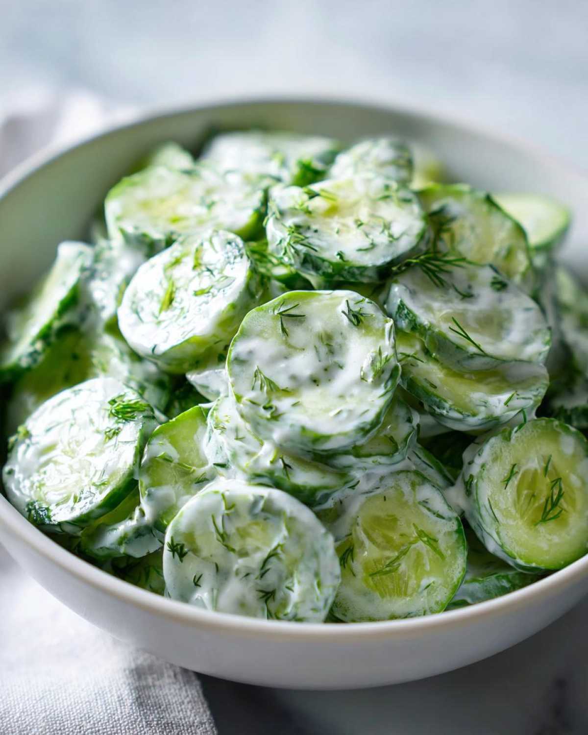 Close-up of a white bowl filled with creamy german cucumber salad, garnished with fresh dill.