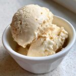 Two scoops of creamy, light-colored protein ice cream in a small white bowl, showing a slightly melted texture.