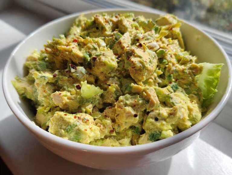 A close-up of a white bowl filled with creamy tuna and avocado salad, garnished with red pepper flakes.