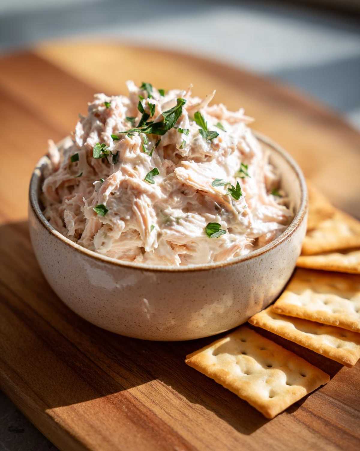 A bowl of creamy tuna dip garnished with fresh parsley, served alongside rectangular crackers on a wooden board.