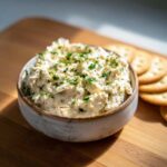A ceramic bowl filled with creamy tuna dip, garnished with fresh chopped parsley, on a wooden cutting board with crackers.