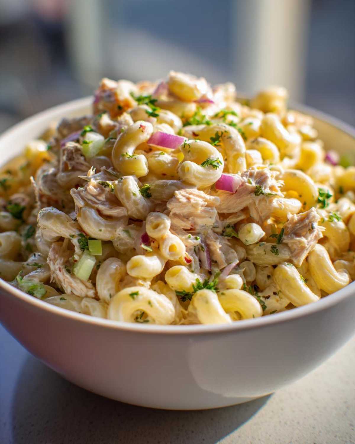 Close-up of a white bowl filled with creamy tuna egg pasta salad, featuring elbow macaroni, tuna, red onion, and parsley.