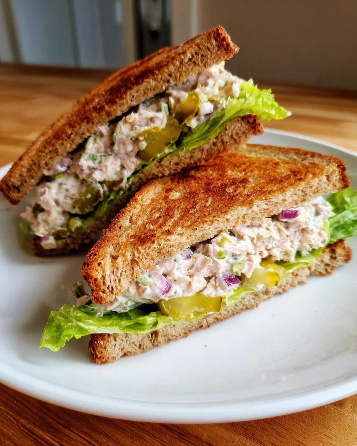 Two halves of a tuna fish sandwich on toasted whole wheat bread with lettuce, red onion, and pickles.