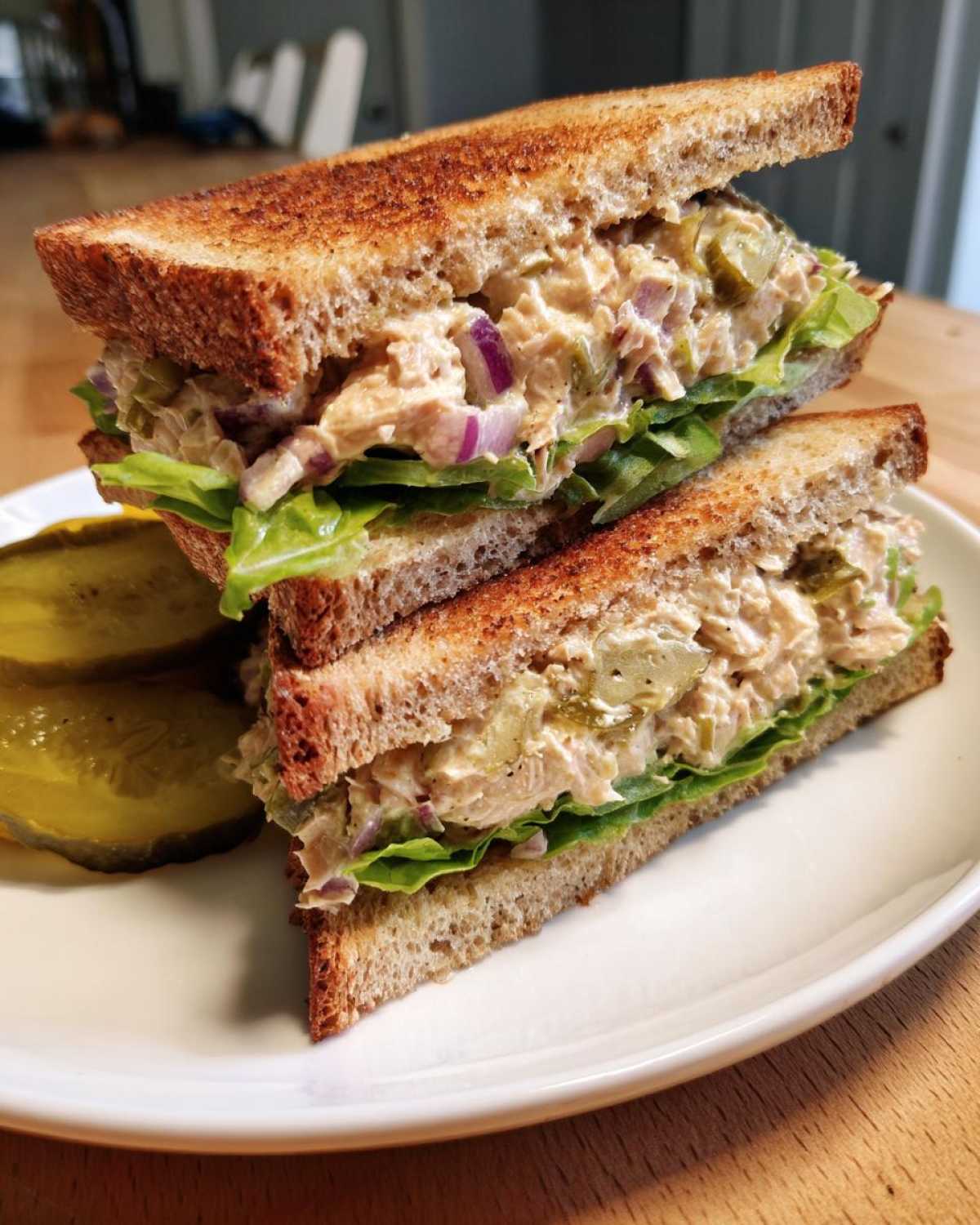A delicious tuna fish sandwich cut in half, revealing creamy tuna salad, lettuce, and red onion, served with pickle slices.