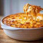 A serving spoon lifting a gooey, cheesy scoop of crockpot buffalo chicken dip from a white baking dish, showing melted cheese pulls.
