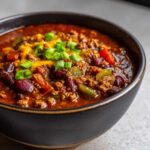 A bowl of hearty crockpot chili topped with cheese and green onions, showcasing the rich texture and ingredients.