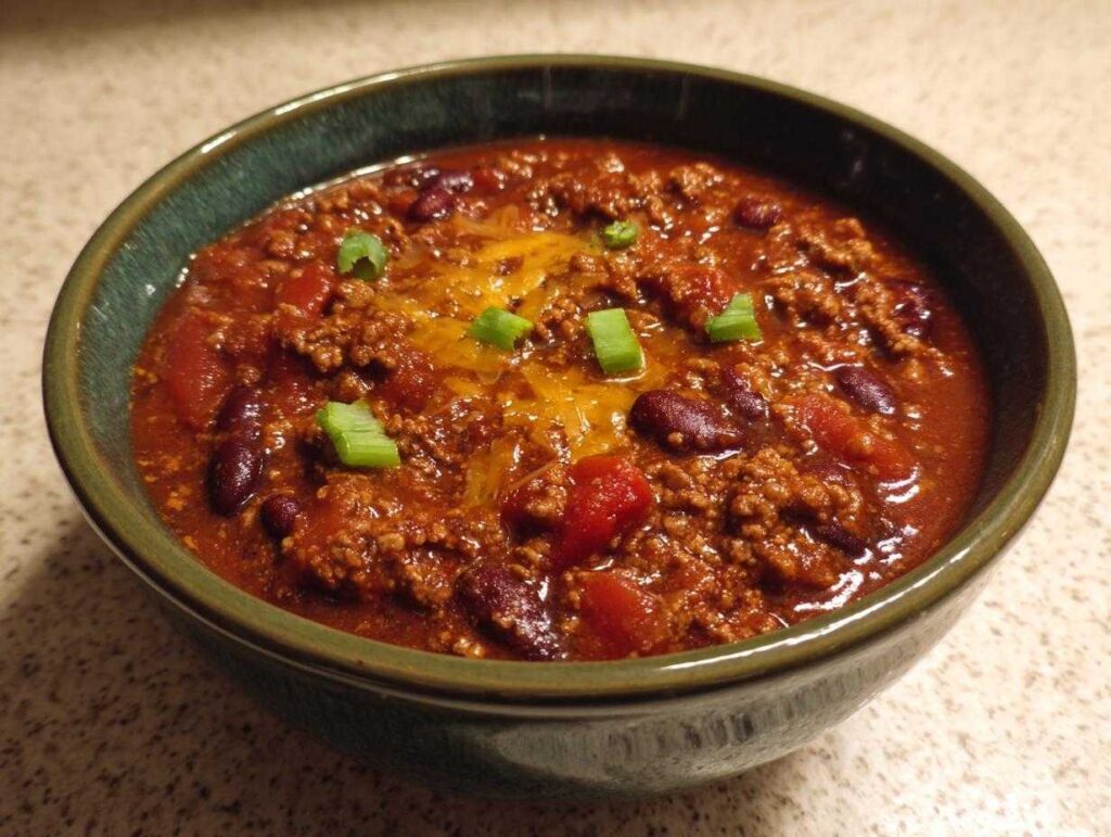A bowl of crockpot chili with kidney beans, tomatoes, ground beef, cheese, and green onion garnish.