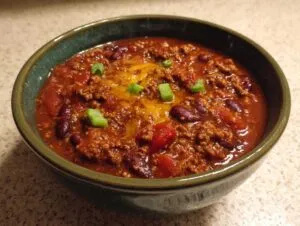 A bowl of crockpot chili with kidney beans, tomatoes, ground beef, cheese, and green onion garnish.