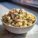 A close-up shot of a white bowl filled with creamy tuna egg pasta salad, garnished with fresh herbs and red onion.