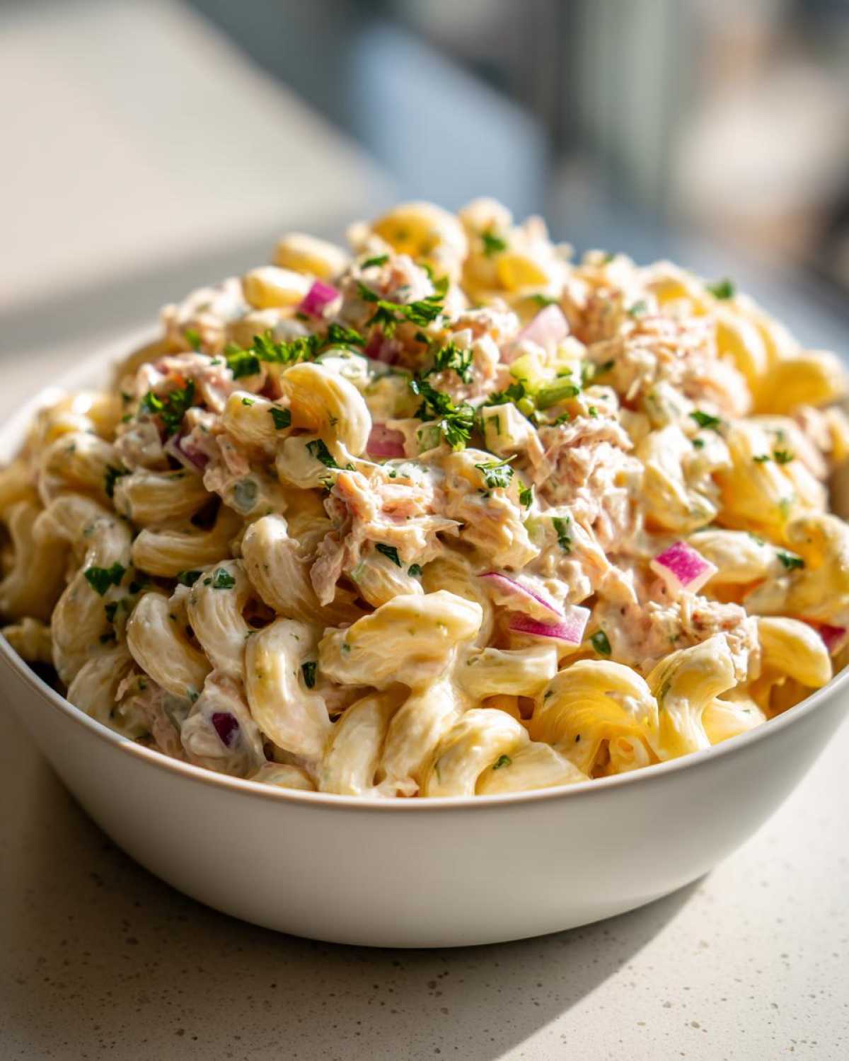 A close-up shot of a large white bowl filled with creamy tuna egg pasta salad, garnished with fresh parsley and red onion.