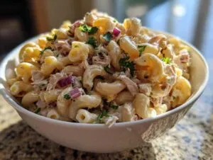 A close-up of a large white bowl filled with creamy tuna egg pasta salad, garnished with fresh parsley and red onion.