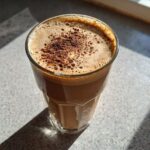 Close-up of a glass of frothy protein coffee topped with a sprinkle of cocoa powder, bathed in sunlight.