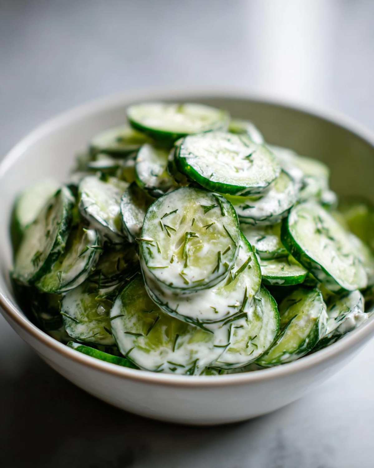 A close-up shot of a bowl filled with creamy german cucumber salad, showing thinly sliced cucumbers coated in a white dressing with dill.