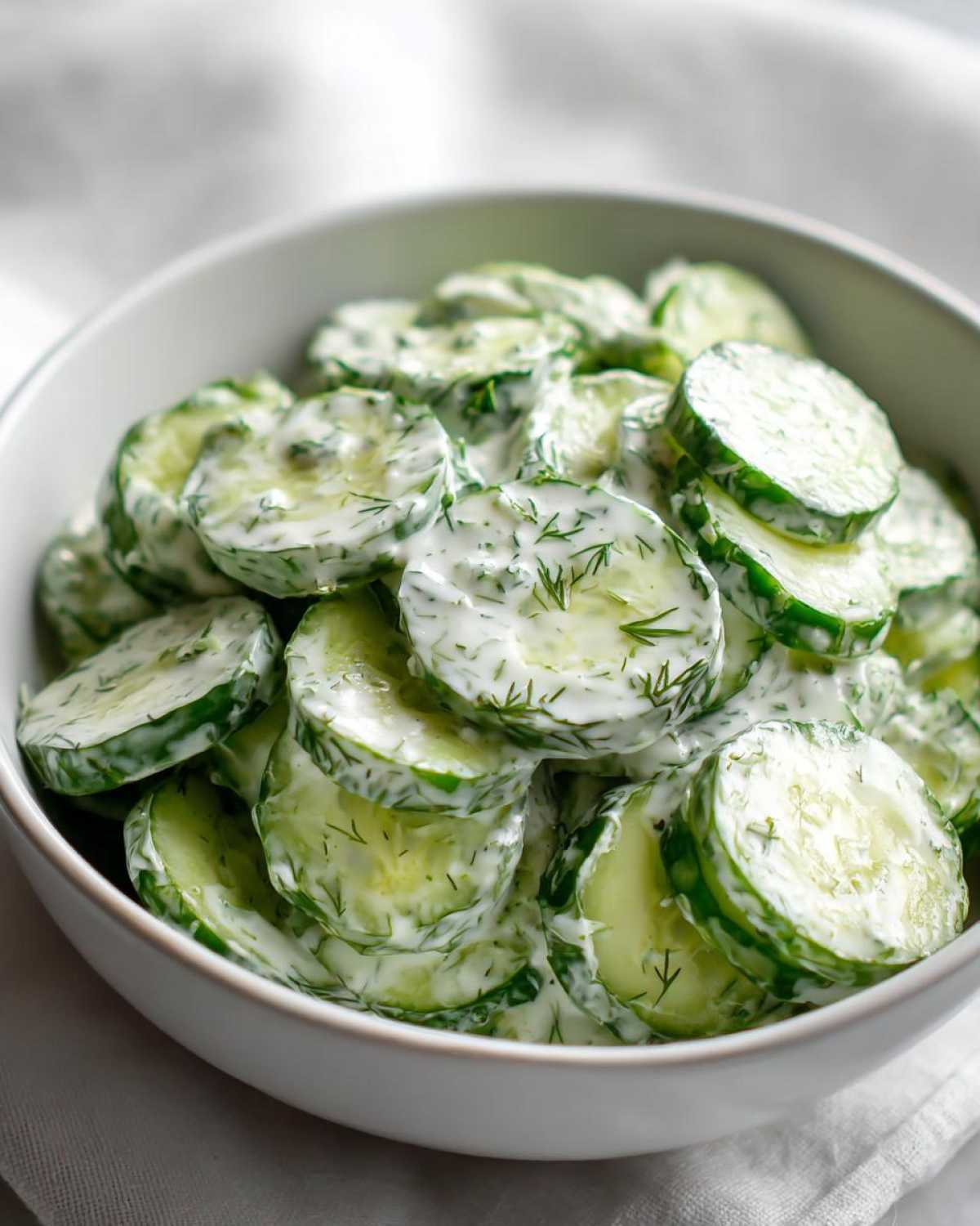 A white bowl filled with creamy german cucumber salad, showing thinly sliced cucumbers coated in a white dressing with dill.