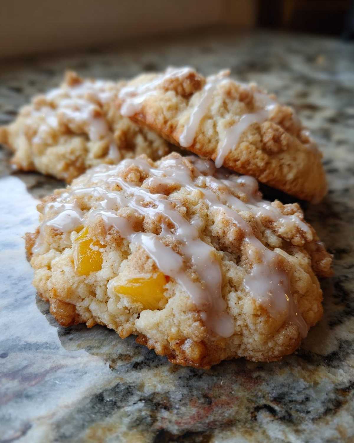 Close-up of two glazed peach cobbler cookies with visible peach pieces and crumb topping on a marble surface.