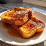Two slices of golden brown protein french toast, drizzled with maple syrup and sprinkled with cinnamon, on a white plate.