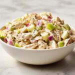 A close-up view of a white bowl filled with high protein chicken salad, featuring shredded chicken, diced red onion, and green apple pieces.
