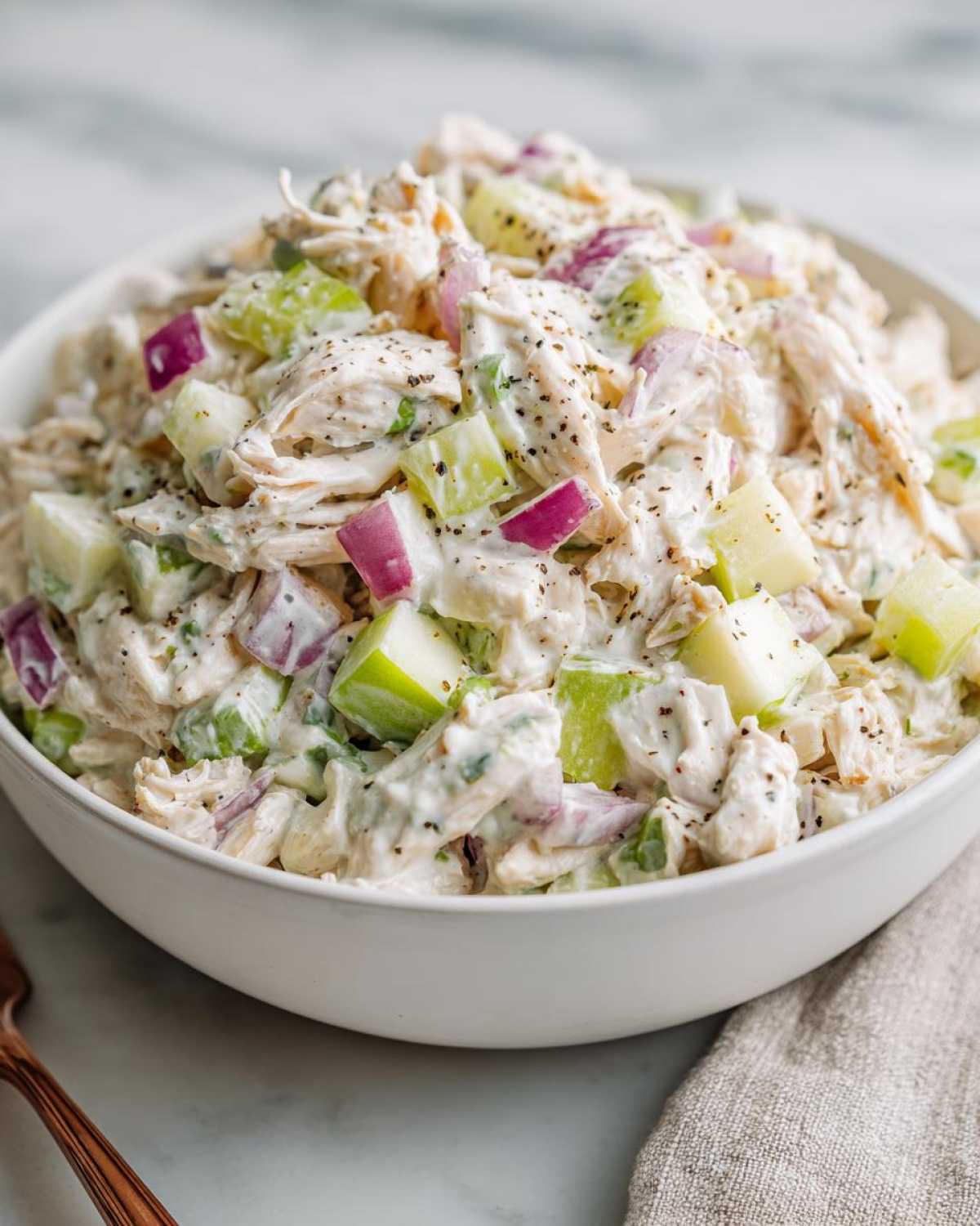 Close-up of a white bowl filled with high protein chicken salad, featuring shredded chicken, red onion, green apple, and celery.