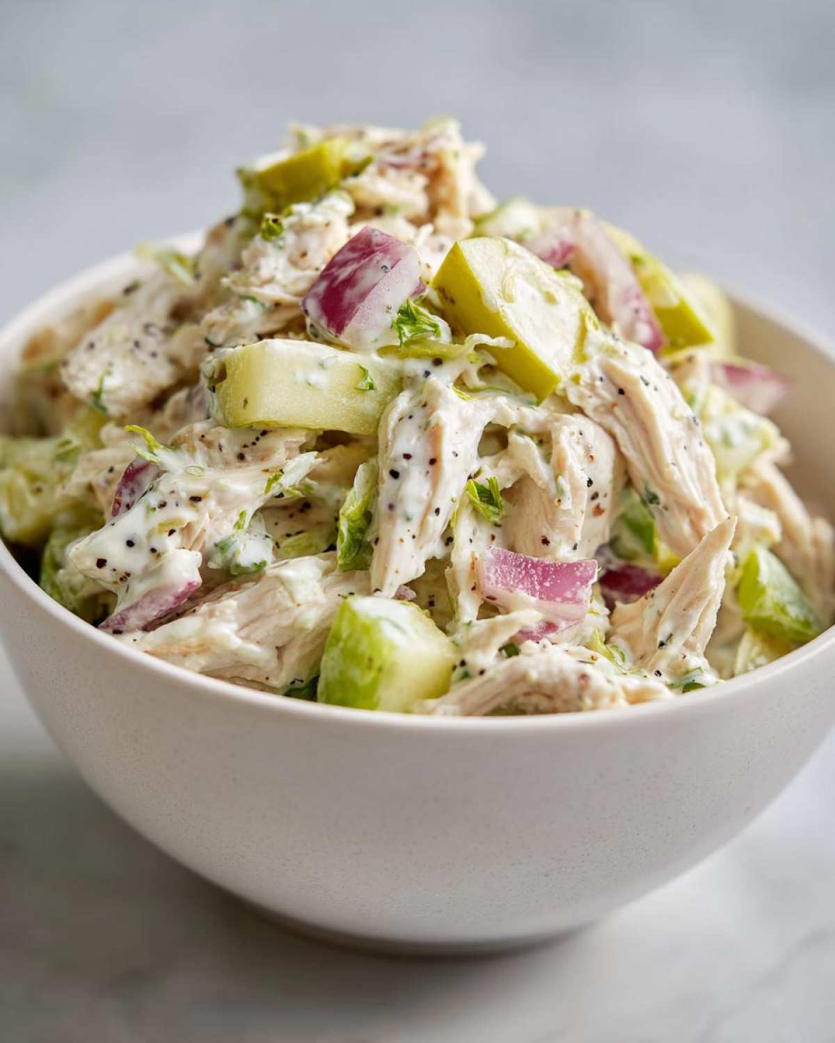 A close-up shot of a white bowl filled with high protein chicken salad, showing shredded chicken, red onion, and green apple pieces.