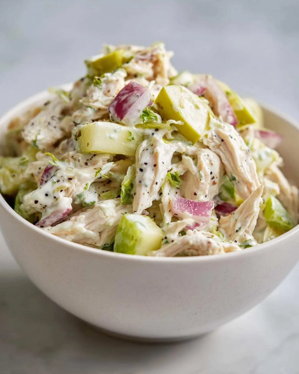 A close-up shot of a white bowl filled with high protein chicken salad, showing shredded chicken, red onion, and green apple pieces.