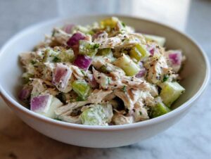 A close-up shot of a white bowl filled with high protein chicken salad, featuring shredded chicken, red onion, and celery.