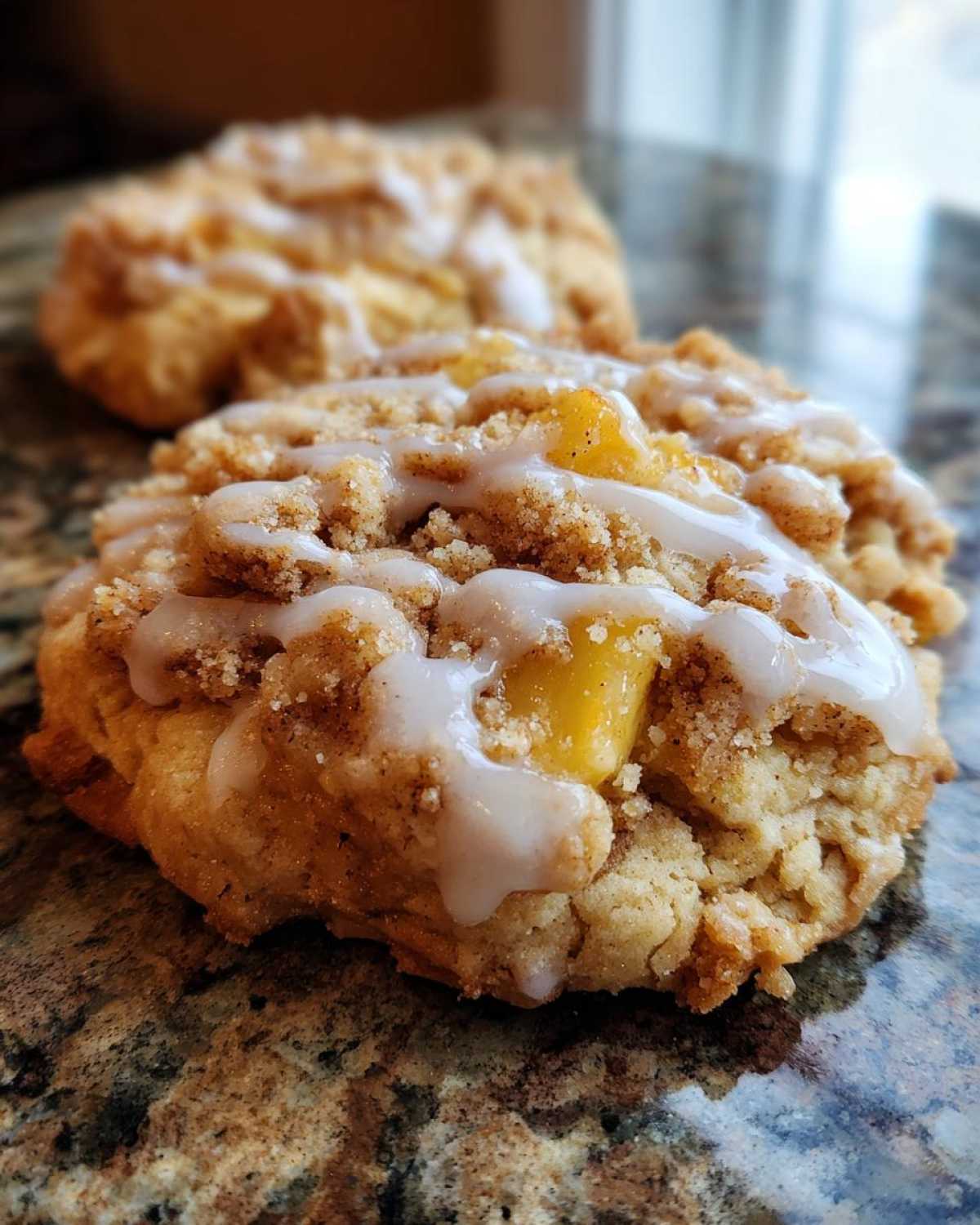 A close-up of a single peach cobbler cookie with a crumb topping and white glaze, sitting on a speckled countertop.