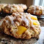 A close-up of a single peach cobbler cookie with a crumbly topping, visible peach pieces, and a drizzle of white icing.