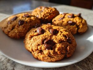 Four golden-brown protein cookies with visible chocolate chips and oatmeal on a white plate, bathed in natural light.