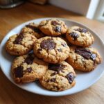 A white plate filled with golden-brown protein chocolate chip cookies, featuring melted chocolate chunks on top.