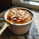 A close-up of a single serving of protein baked oats in a white ramekin, topped with caramel sauce and rolled oats.