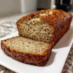 A golden-brown loaf of protein banana bread with a single slice cut and placed in front, showing moist texture.
