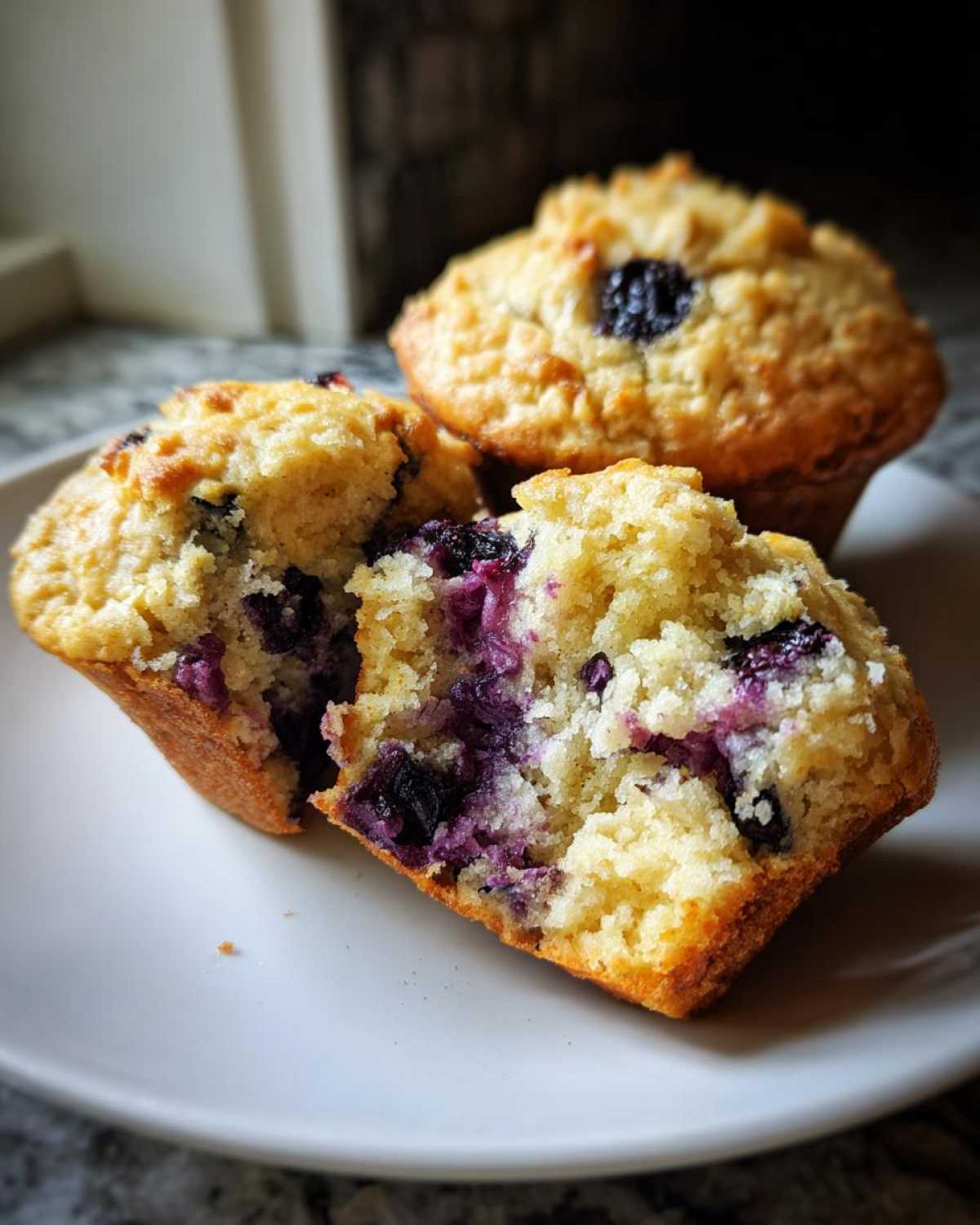 Close-up of three golden-brown protein blueberry muffins on a white plate, one split open revealing juicy blueberries.