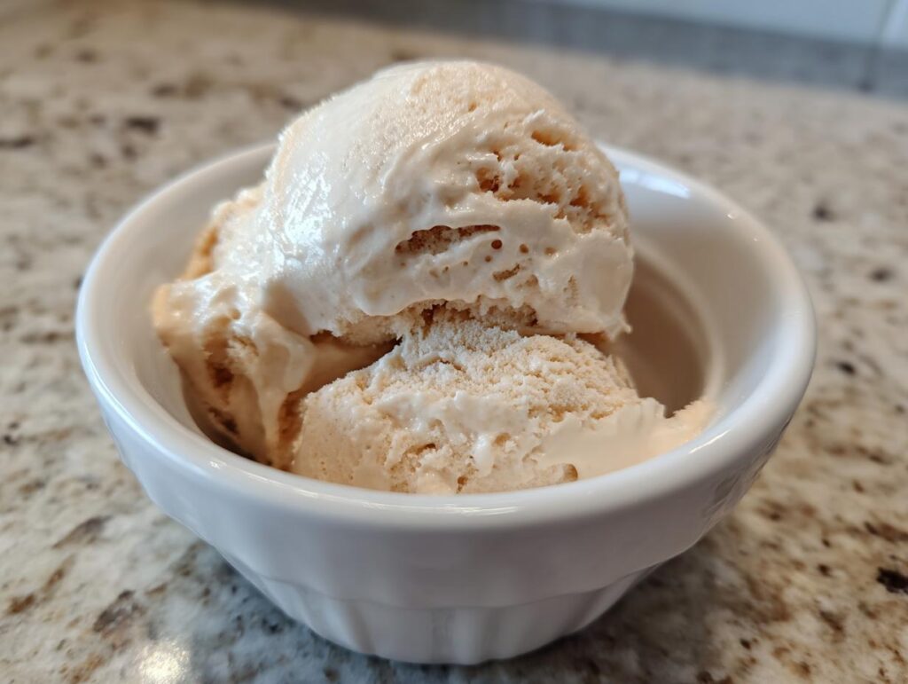 Two scoops of light brown protein ice cream in a white bowl on a speckled countertop.