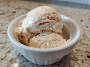 Two scoops of light brown protein ice cream in a white bowl on a speckled countertop.