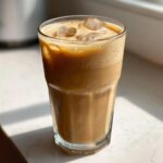A tall glass of protein iced coffee with ice cubes and a foamy top, sitting on a sunlit surface.