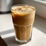 A tall glass of protein iced coffee with ice cubes and a foamy top, sitting on a sunlit surface.