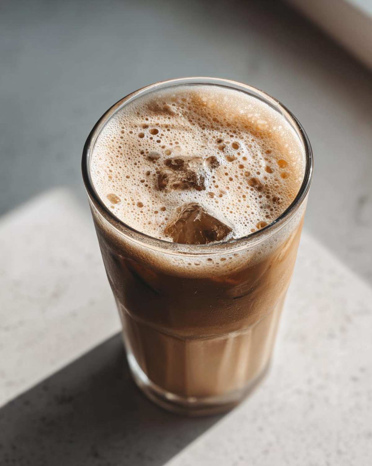 Close-up of a tall glass of protein iced coffee with ice cubes and a foamy top, casting a shadow.