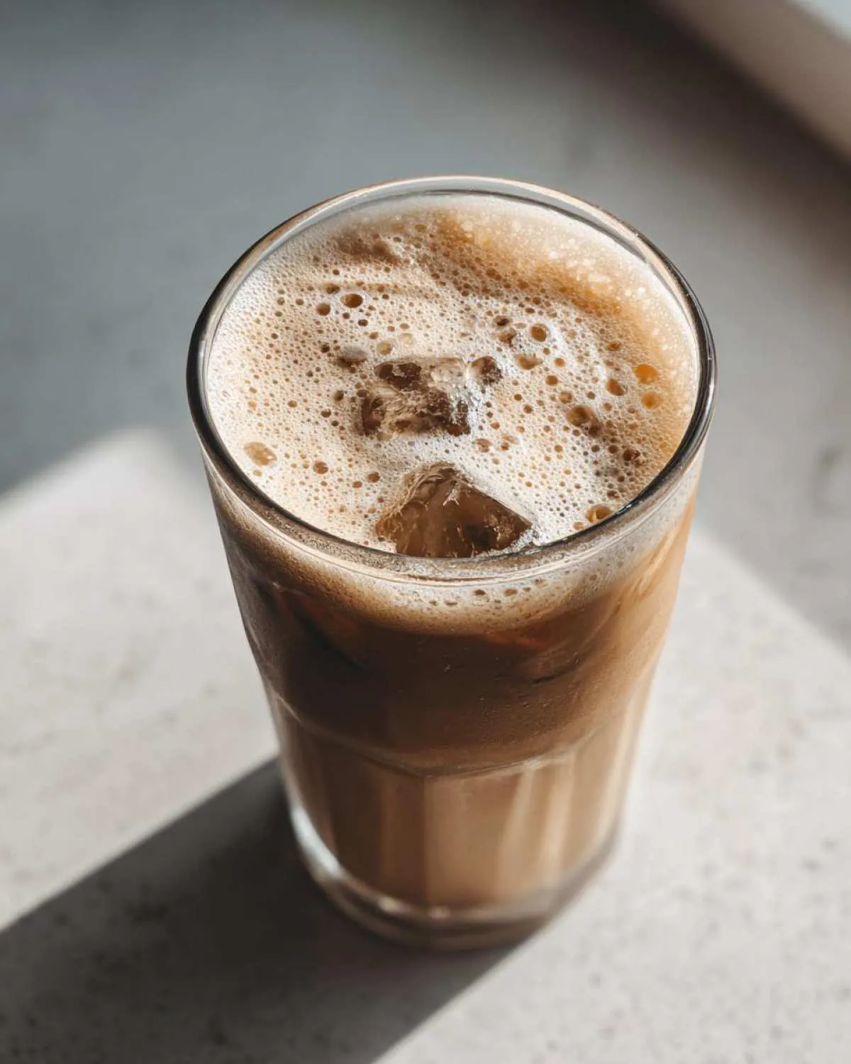 Close-up of a tall glass of protein iced coffee with ice cubes and a foamy top, casting a shadow.
