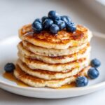 A close-up shot of a stack of golden-brown protein pancakes, topped with fresh blueberries and drizzled with maple syrup.