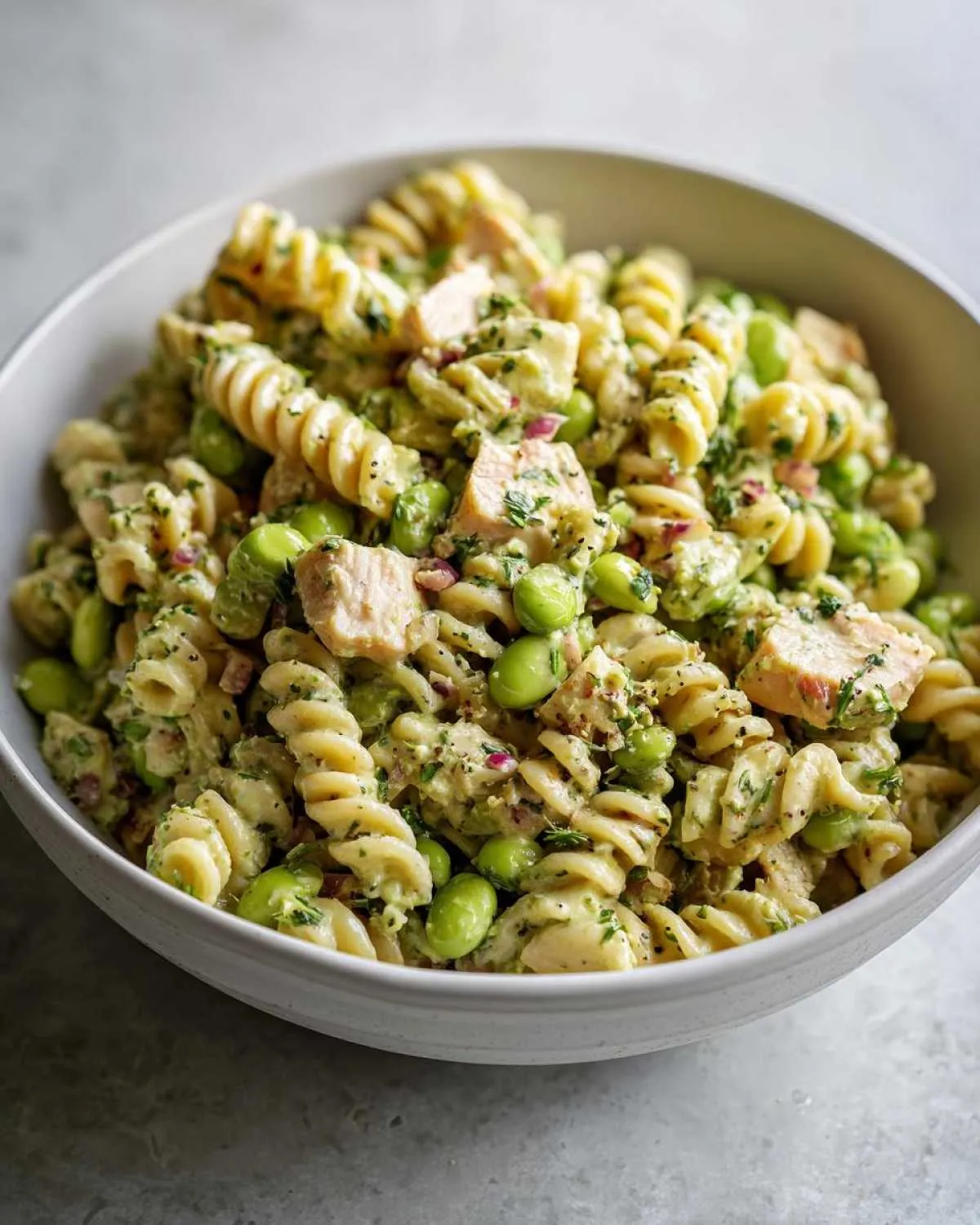 Close-up of a bowl filled with protein pasta salad, featuring rotini pasta, edamame, chicken pieces, and herbs.