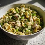 A close-up of a bowl filled with protein pasta salad, featuring orecchiette pasta, edamame, and shredded chicken or tuna, coated in a green dressing and garnished with herbs.