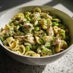 A close-up of a bowl filled with protein pasta salad, featuring orecchiette pasta, edamame, and shredded chicken or tuna, coated in a green dressing and garnished with herbs.