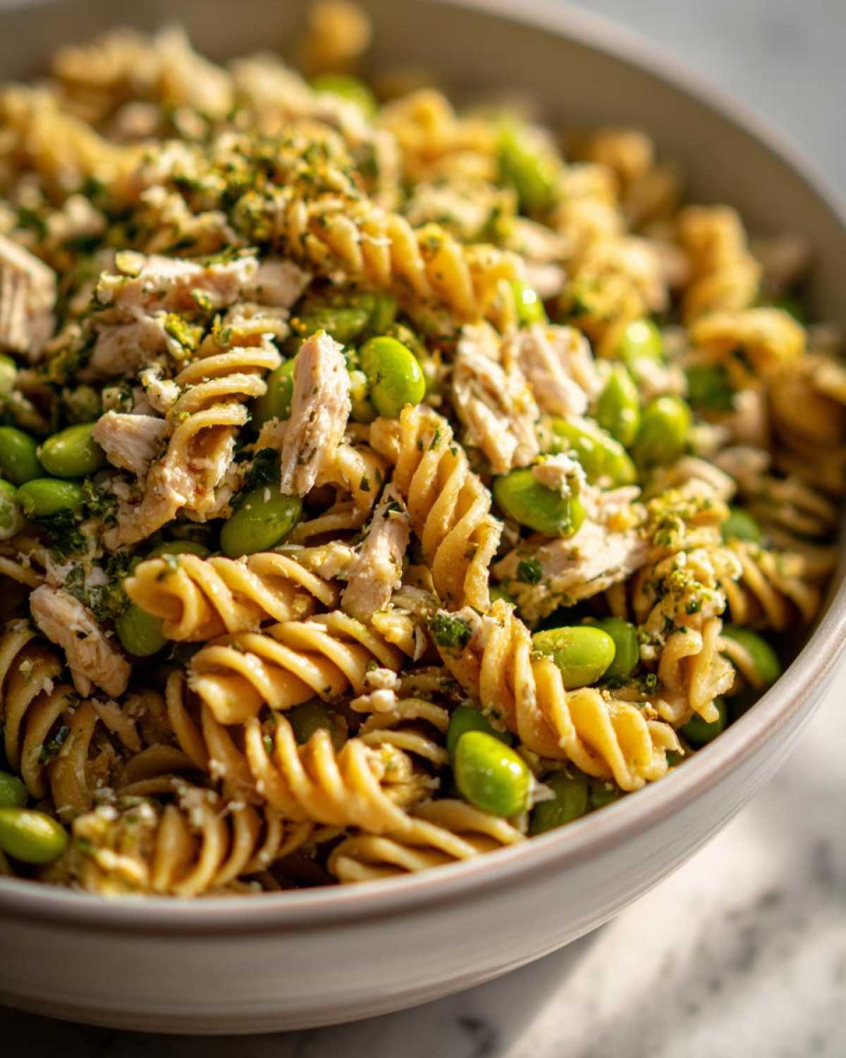 Close-up of a bowl filled with protein pasta salad, featuring spiral pasta, shredded chicken, bright green edamame, and herbs.