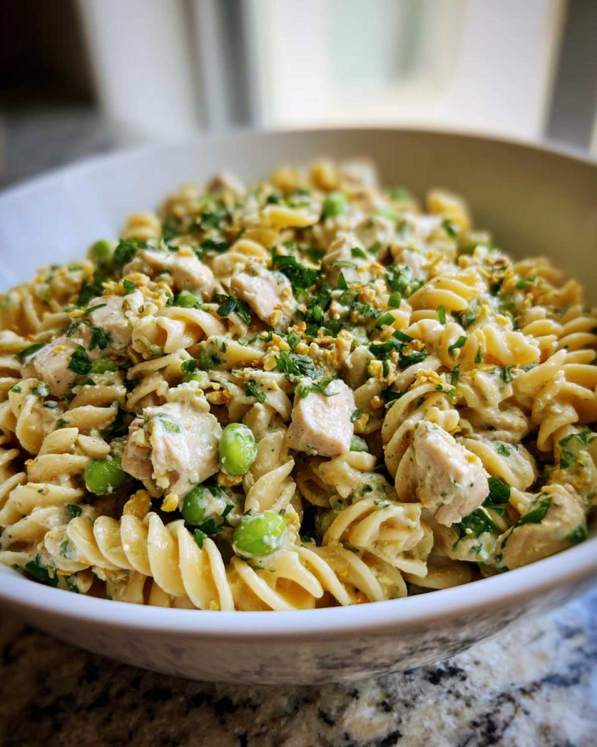 Close-up of a bowl filled with protein pasta salad, featuring spiral pasta, diced chicken, green peas, and fresh herbs.
