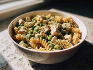 A close-up of a bowl of protein pasta salad with rotini pasta, diced chicken, green peas, and a creamy dressing.