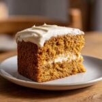 A slice of pumpkin spice cake with white frosting on a white plate, showcasing the moist texture.