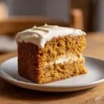 A slice of pumpkin spice cake with white frosting on a white plate, showcasing the moist texture.