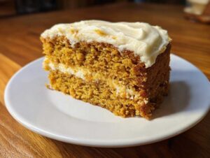 A slice of moist pumpkin spice cake with cream cheese frosting on a white plate, ready to eat.